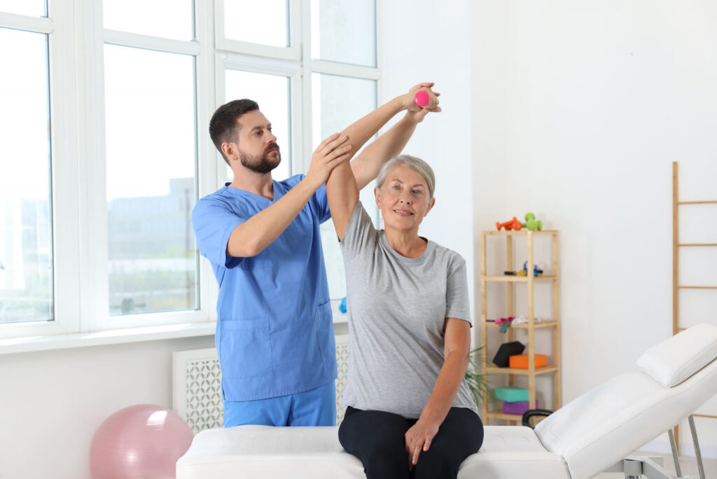 Physical therapist guiding a senior woman through a shoulder rehabilitation exercise in a bright clinic