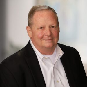 Corporate headshot of a man in a black blazer and white shirt against a softly blurred office background