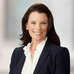 Corporate headshot of a woman in a navy blazer against a bright, softly blurred office