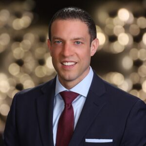 Executive headshot of a man in a navy suit and red tie with warm background lights